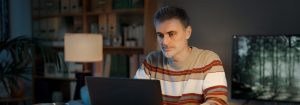 Een man met een gestreepte trui zit aan zijn bureau en kijkt naar de laptop voor hem.