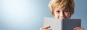 "Jong kind met blond haar glimlacht achter open boek, toont vreugdevolle leeservaring tegen lichtblauwe achtergrond."